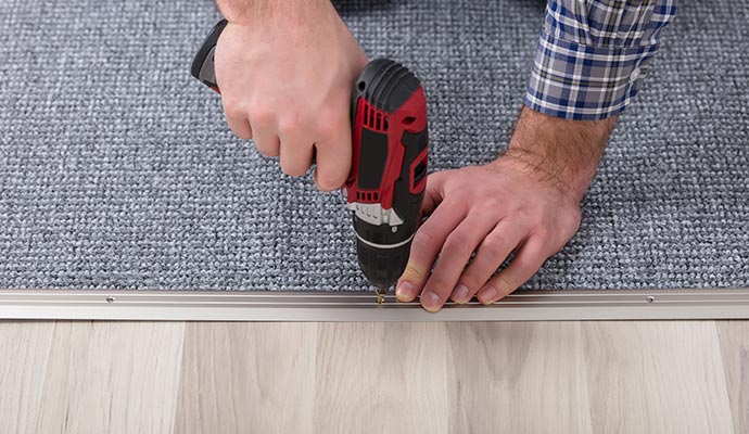 An expert securing metal tack strip on gray carpet with power drill