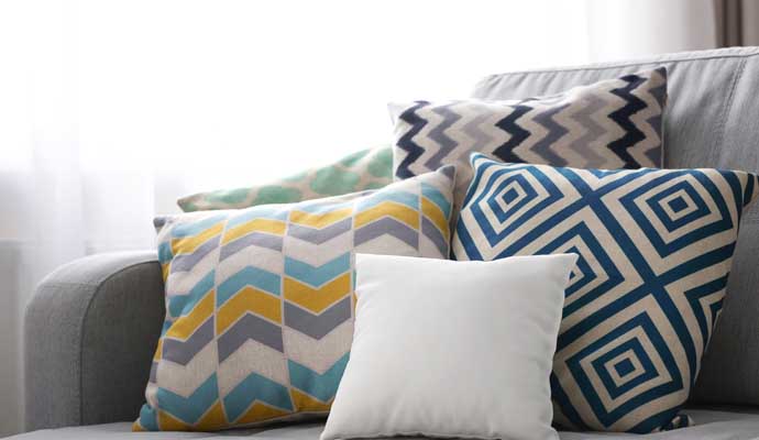 A stack of colorful, geometric-patterned throw pillows and one plain white cushion arranged on a grey sofa